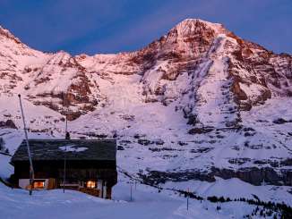 The Eiger and the Mönch tower over a snow-covered landscape as twilight begins to fall. Warm light illuminates individual buildings, while the rocky and snow-covered slopes of the mountains glow against the evening sky.