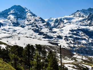 The Wengernalpbahn runs through alpine terrain under a bright sun. The Eiger, Mönch, and Jungfrau dominate the background. 