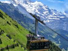 The Wengen–Männlichen aerial tramway glides uphill over steep alpine meadows. In the background, the impressive high-mountain landscape rises up, highlighting the tramway’s spectacular location.