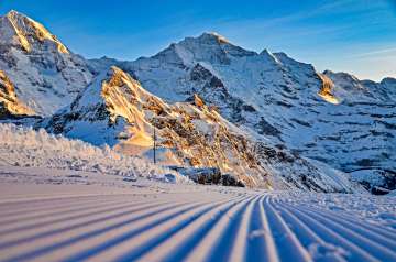 A freshly groomed ski slope with distinct tracks runs through the foreground, winding its way through the snow-covered high mountains. In the warm morning light, the rocky and snow-covered slopes of the Eiger and Mönch glow in the background.