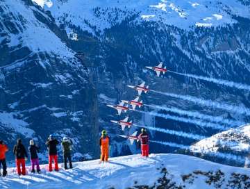 Planes from the Patrouille Suisse fly in tight formation over the Lauberhorn Race. In the foreground, spectators stand on a snow-covered slope watching the air show, while the planes leave trails of smoke against the steep, snow-covered mountainside.