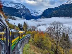 The Wengernalpbahn runs along the mountainside above the Lauterbrunnen Valley. Fog hangs in the valley, while forests and steep cliffs line the route.