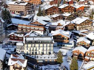 Aerial view of the mountain village of Wengen with snow-covered chalets, hotels, and streets. The village is nestled on the hillside below a wintry forest.