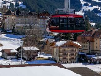 A gondola on the Wengen–Männlichen cable car floats above the snow-covered village of Wengen. Sports facilities and buildings can be seen below, while the mountain backdrop frames the village in the distance.