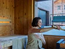 Two guests in the sauna at the Hotel Fiescherblick in Grindelwald, looking out through the panoramic window at the hotel terrace and the surrounding buildings.