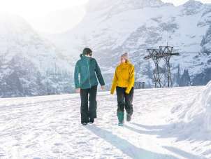 Two people are walking from the First mountain station toward Oberjoch along a snow-covered winter hiking trail. The low-hanging sun creates a bright backlight and long shadows in the snow. Distinctive, snow-capped mountain peaks are visible in the background.