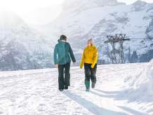 Two people are walking from the First mountain station toward Oberjoch along a snow-covered winter hiking trail. The low-hanging sun creates a bright backlight and long shadows in the snow. Distinctive, snow-capped mountain peaks are visible in the background.