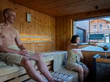 Two guests in the sauna at the Hotel Fiescherblick in Grindelwald, looking out through the panoramic window at the hotel terrace and the surrounding buildings.