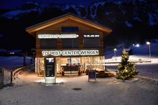 The Tourist Center is brightly lit on a winter evening in Wengen. An illuminated Christmas tree stands in front of the building.