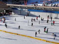 The large-scale ice rink in Wengen is bustling with skaters. Children and adults glide across the ice, while spectators stand at the edge, getting ready. In the background, village buildings and other winter sports facilities are visible.
