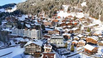Aerial view of the mountain village of Wengen with snow-covered chalets, hotels, and streets. The village is nestled on the hillside below a wintry forest.
