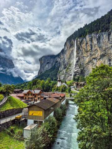 A view of the village of Lauterbrunnen with its chalets and the flowing Lütschine River. In the background, the Staubbach Falls plunge from the steep rock face into the valley, defining the village’s skyline.