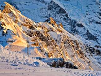 A freshly groomed ski slope with distinct tracks runs through the foreground, winding its way through the snow-covered high mountains. In the warm morning light, the rocky and snow-covered slopes of the Eiger and Mönch glow in the background.