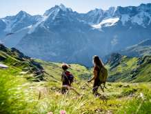 Two friends are hiking in Blumental in Mürren. They are enjoying the view of the green landscape with the white mountain panorama—featuring the Eiger, Mönch, and Jungfrau—in the background.