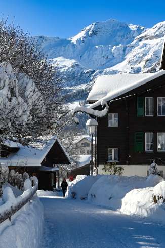 A snow-covered village path in Wengen, winding between traditional chalets. In the background, the Jungfrau rises with its snow-capped slopes under a clear sky.