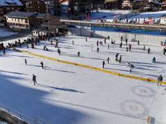 The large ice rink in Wengen is bustling with skaters. Children and adults glide across the ice, while spectators stand at the edge, getting ready. In the background, you can see the village buildings and other winter sports facilities.