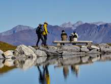 Two people are standing on a stone path by Lake Fallboden near Kleine Scheidegg. The surrounding mountain landscape is reflected in the clear water; the scene appears peaceful and expansive.